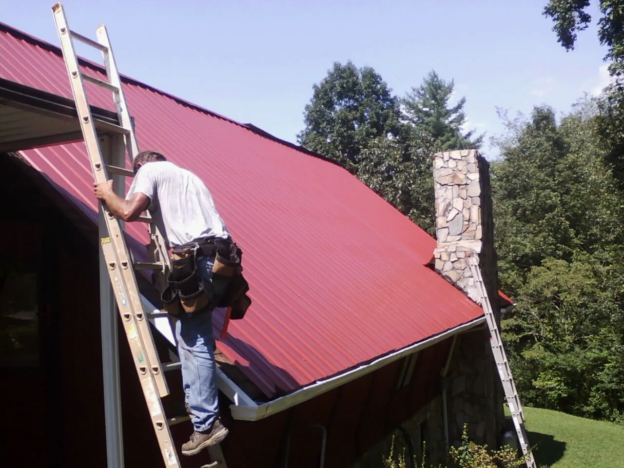 Roofer on a ladder beside a red metal roof and stone chimney — Soffit & Fascia Repair by Roofing Brothers in Silver Creek